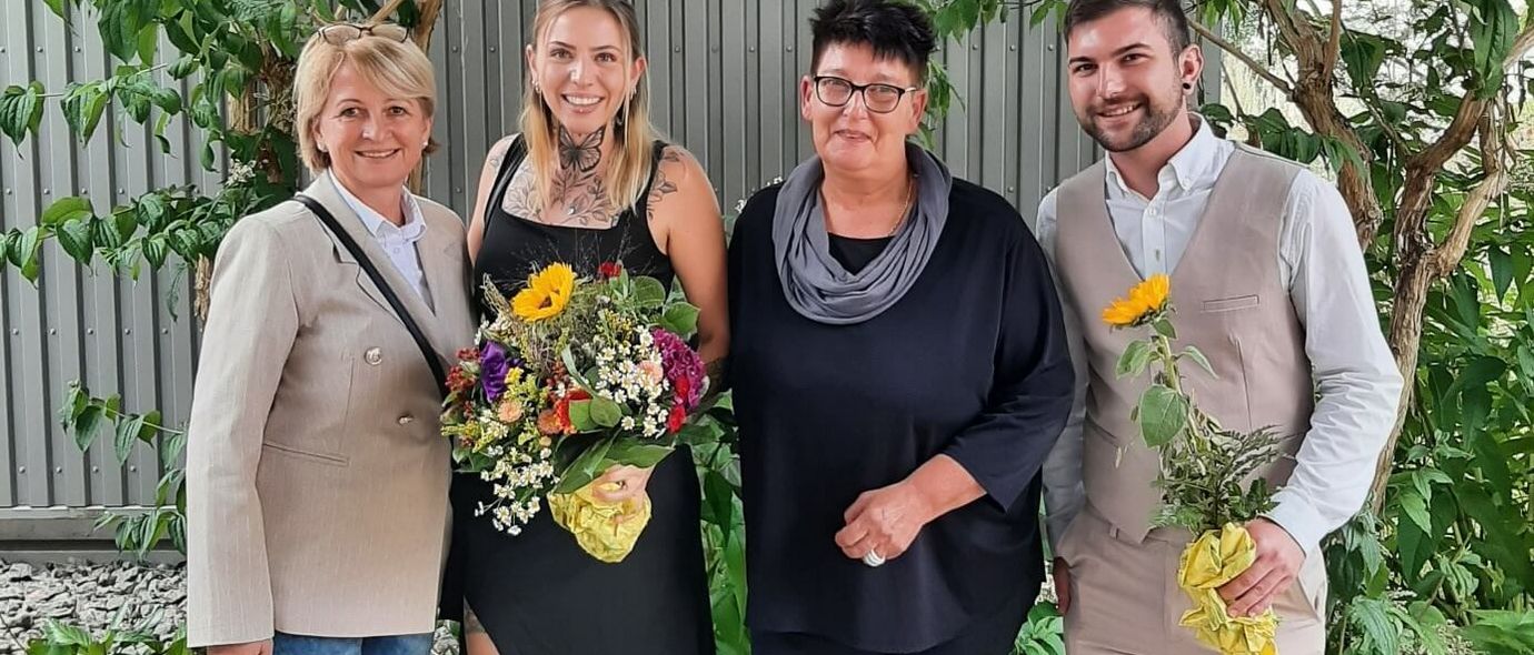 Auszubildende und Ausbilder stehen mit Blumen vor einem Zaun