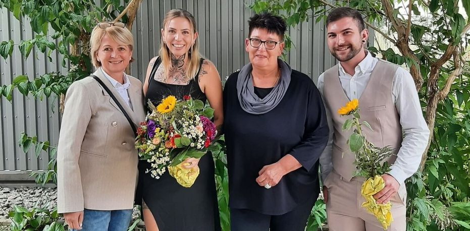 v. l. n. r.: Hida Jusic (PDL und Praxisanleiterin), Anna-Lena Maier, Eike Fundinger (HL), David Goger Auszubildende und Ausbilder stehen mit Blumen vor einem Zaun
