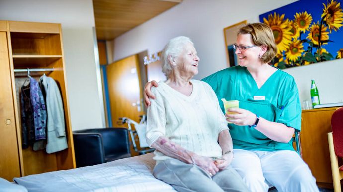 Pflegekraft reicht Seniorin einen Becher mit Saft, während Sie auf dem Bett sitzen